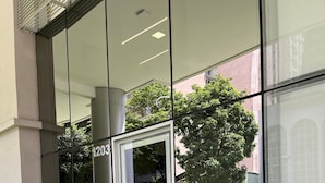 Entrada de un edificio moderno con puerta de vidrio, letrero con el número '1203' y un pequeño espacio exterior con plantas.