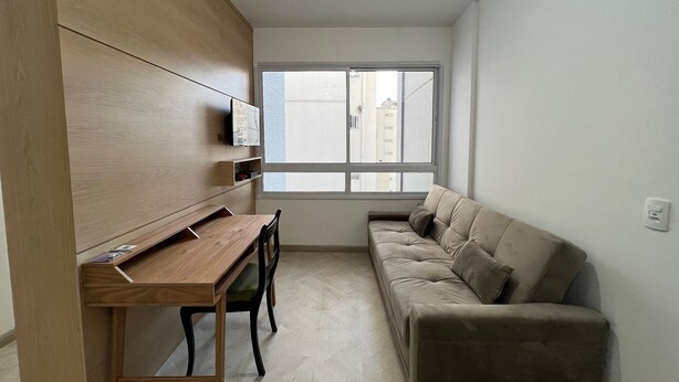 A small, modern room with a wooden desk, a cushioned sofa, and a large window. - LK Centro 9 (Belo Horizonte)