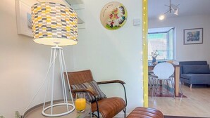 A living room with a lamp, a sofa, an ottoman, a mirror with lights, a round wall decoration, and a dining area in the background. - NEW Family Home in Whitstable | Pets Welcome (Whitstable)