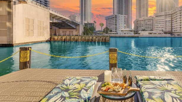 A waterfront dining area with a view of a city skyline at sunset, featuring high-rise buildings and a dock. - 1BR, Marina & Bay View, Resort & Beach (Hallandale Beach)