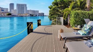 A waterfront lounge area with lounge chairs, a pool, and a city skyline in the background. - 1BR, Marina & Bay View, Resort & Beach (Hallandale Beach)