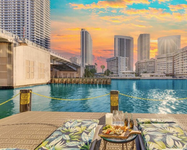 A waterfront view with high-rise buildings, a dock, and a dining setup with a plate of food and drinks. - Cozy Studio w/ Intracoastal View & Balcony (Hallandale Beach)