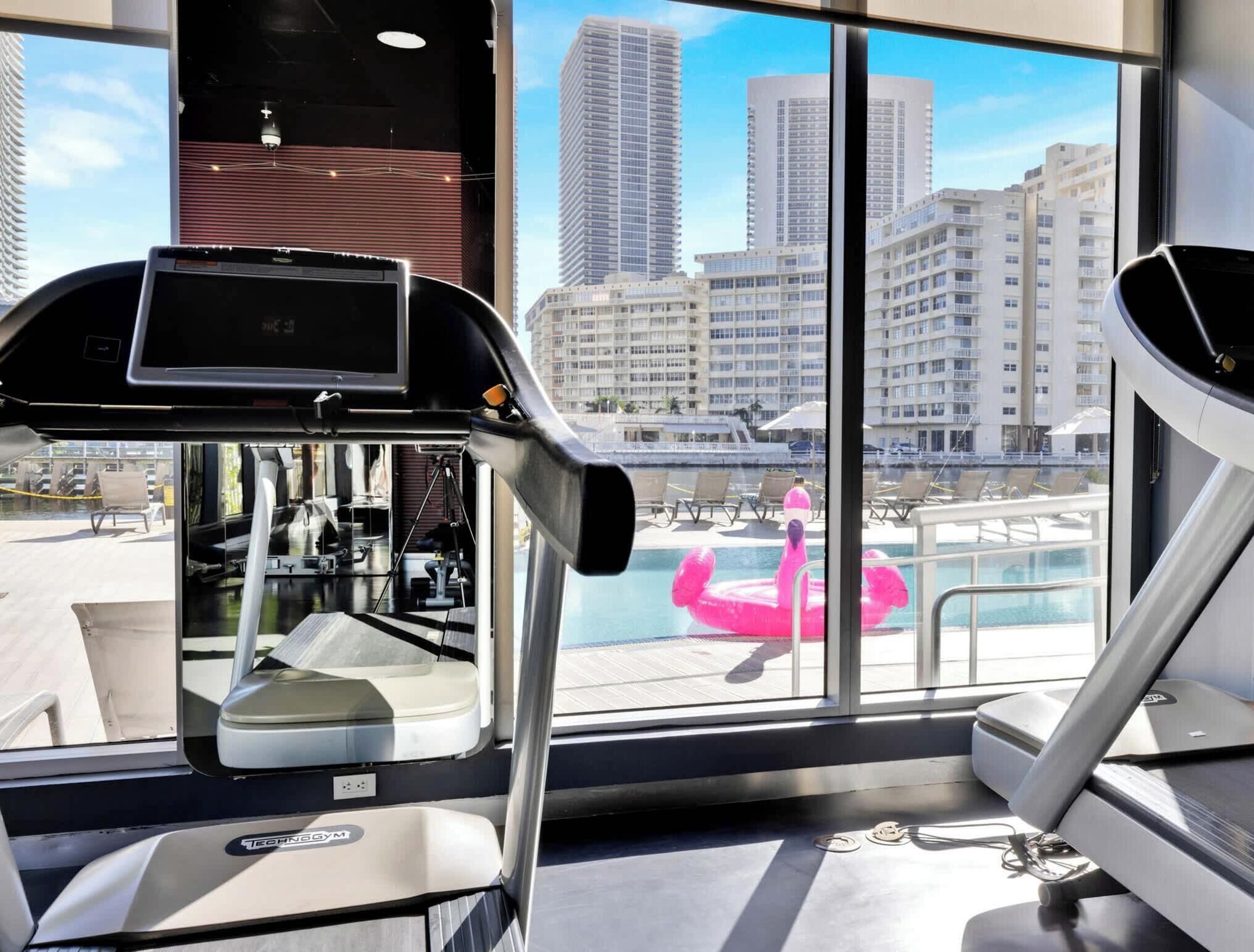 Une salle de sport équipée de matériel d’exercice et offrant une vue sur le paysage urbain grâce à de grandes fenêtres.