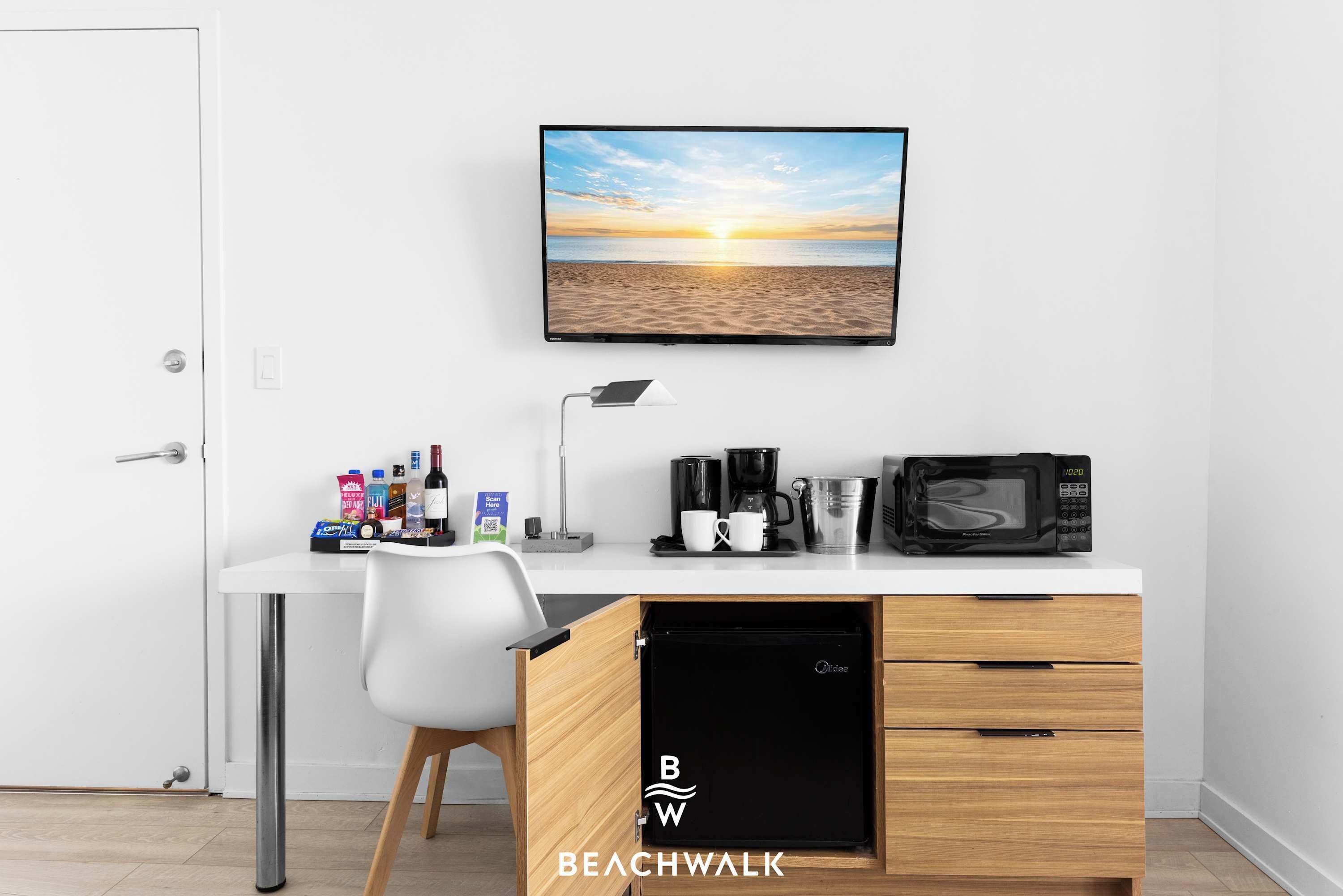 Une chambre d’hôtel moderne avec un bureau blanc, un téléviseur fixé au mur diffusant une scène de plage, une machine à café, un four à micro-ondes et un mini-bar.
