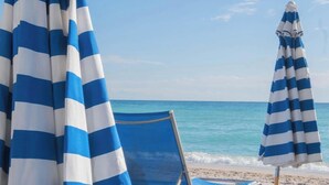 A beach scene with a chair and two striped umbrellas. - 1BR · Sunset View · High Floor · W/D · Resort (Hallandale Beach)