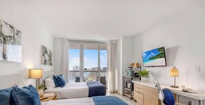 Ein modernes Hotelzimmer mit Bett, Nachttisch, Lampe und Blick auf die Stadt durch große Fenster.