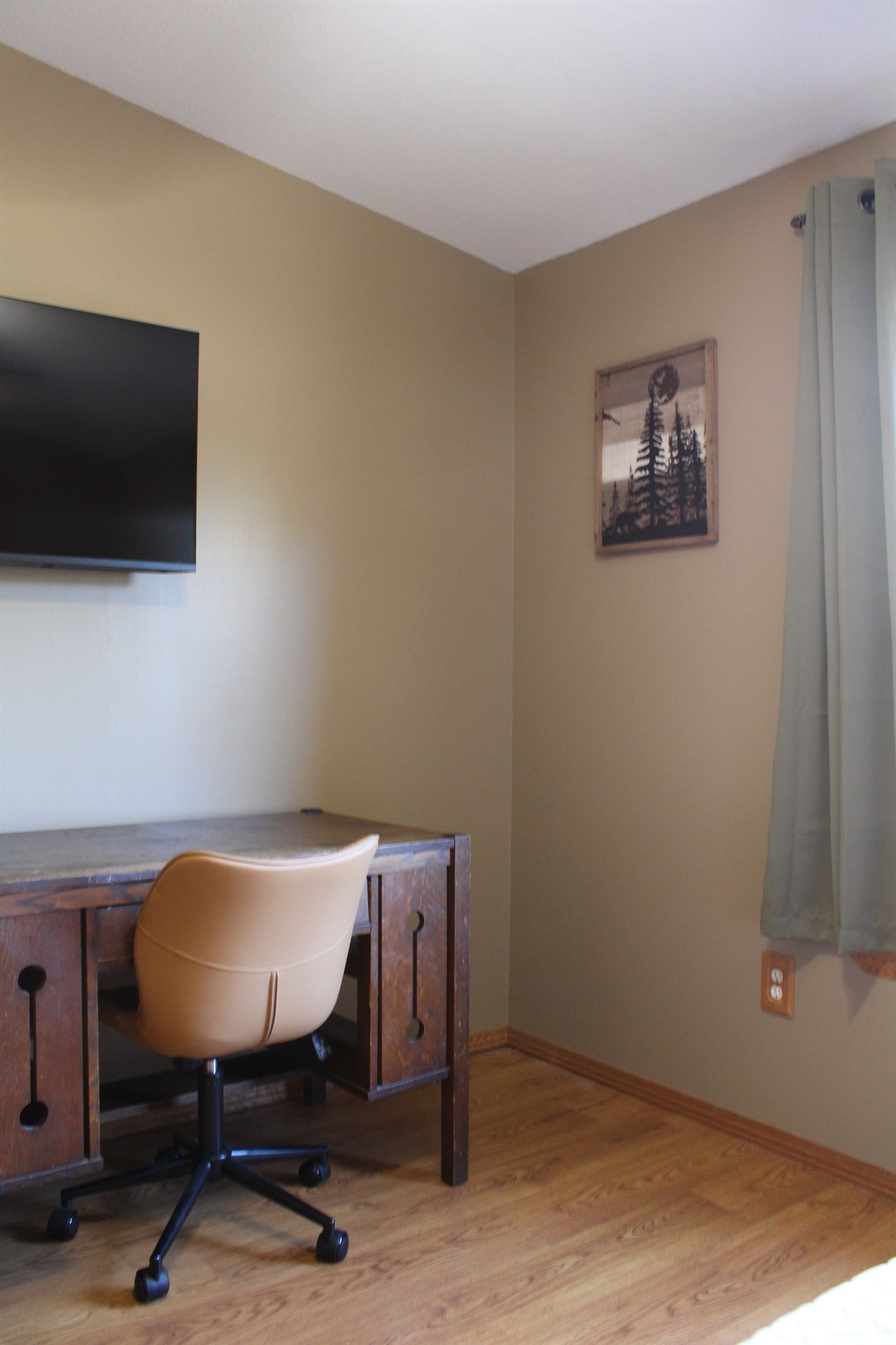 A room with a wooden desk, a chair, a flat-screen TV mounted on the wall, and a framed picture.