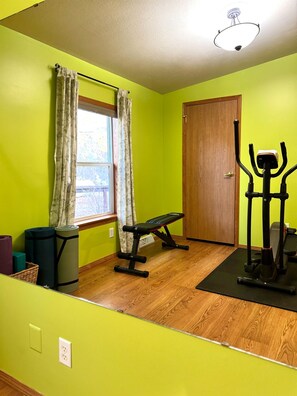 A room with a wooden floor, a window with curtains, a door, and exercise equipment including a bench and a stationary bike.