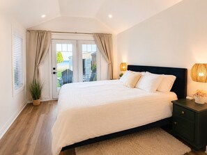 Ein Schlafzimmer mit einem Bett, einem Nachttisch, einer Lampe, einem Blumentopf und Blick ins Freie.