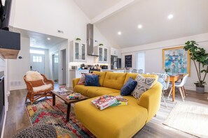 Ein modernes Wohnzimmer mit einem gelben Sofa, einem Essbereich und einer Küche mit weißen Schränken und Edelstahlgeräten.