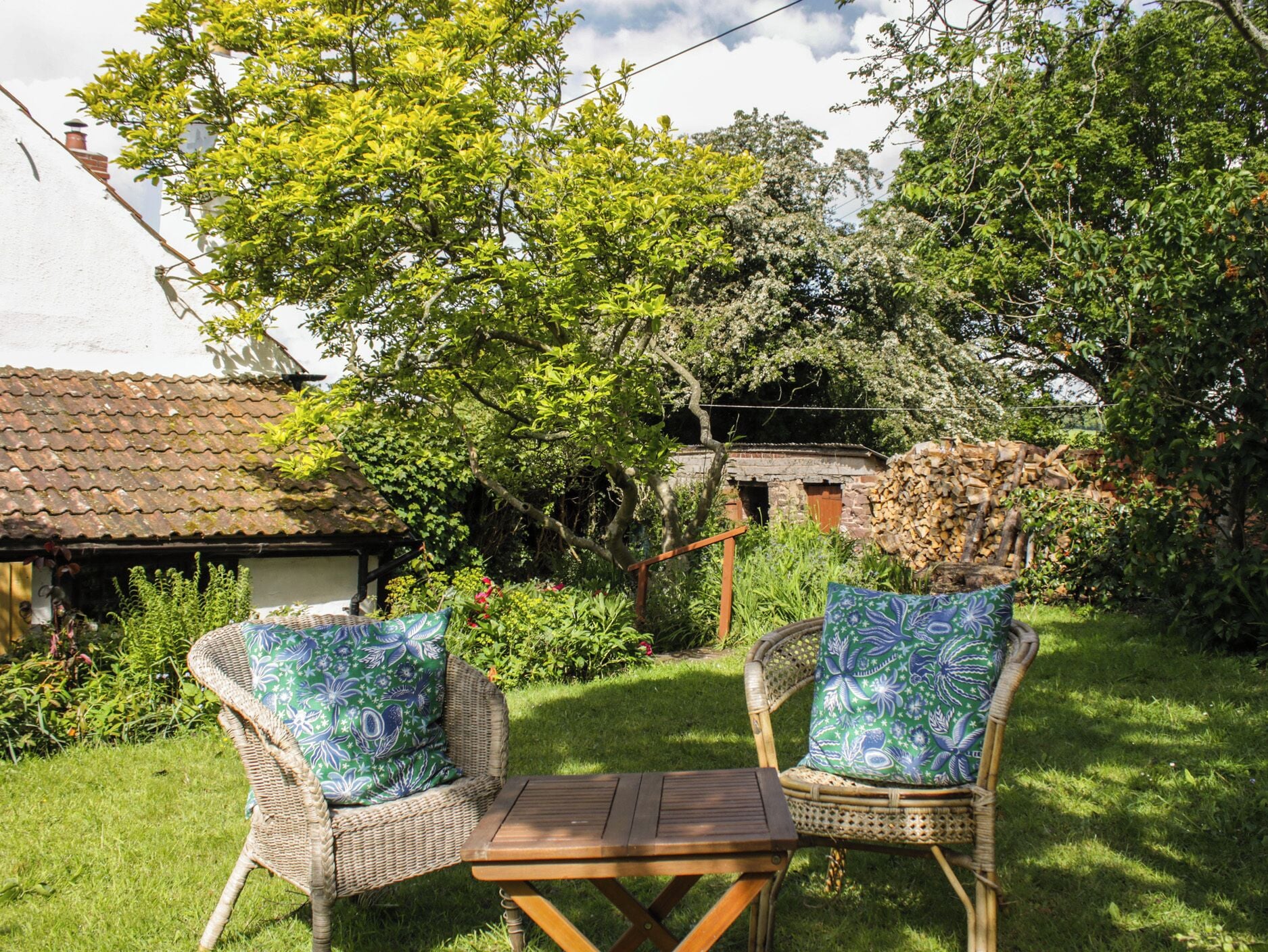 An outdoor seating area with wicker chairs and a wooden table, surrounded by lush greenery and a rustic building.