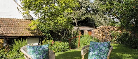 An outdoor seating area with wicker chairs and a wooden table, surrounded by lush greenery and a rustic building.