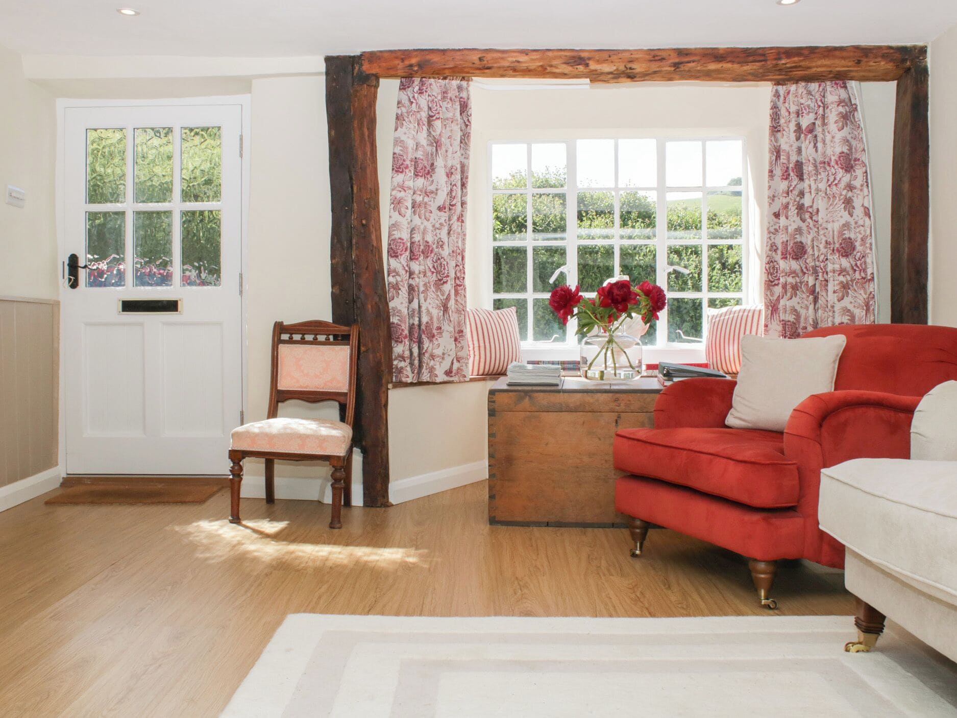 A room with a wooden door, a window with floral curtains, a red sofa, a wooden chest, and a chair.