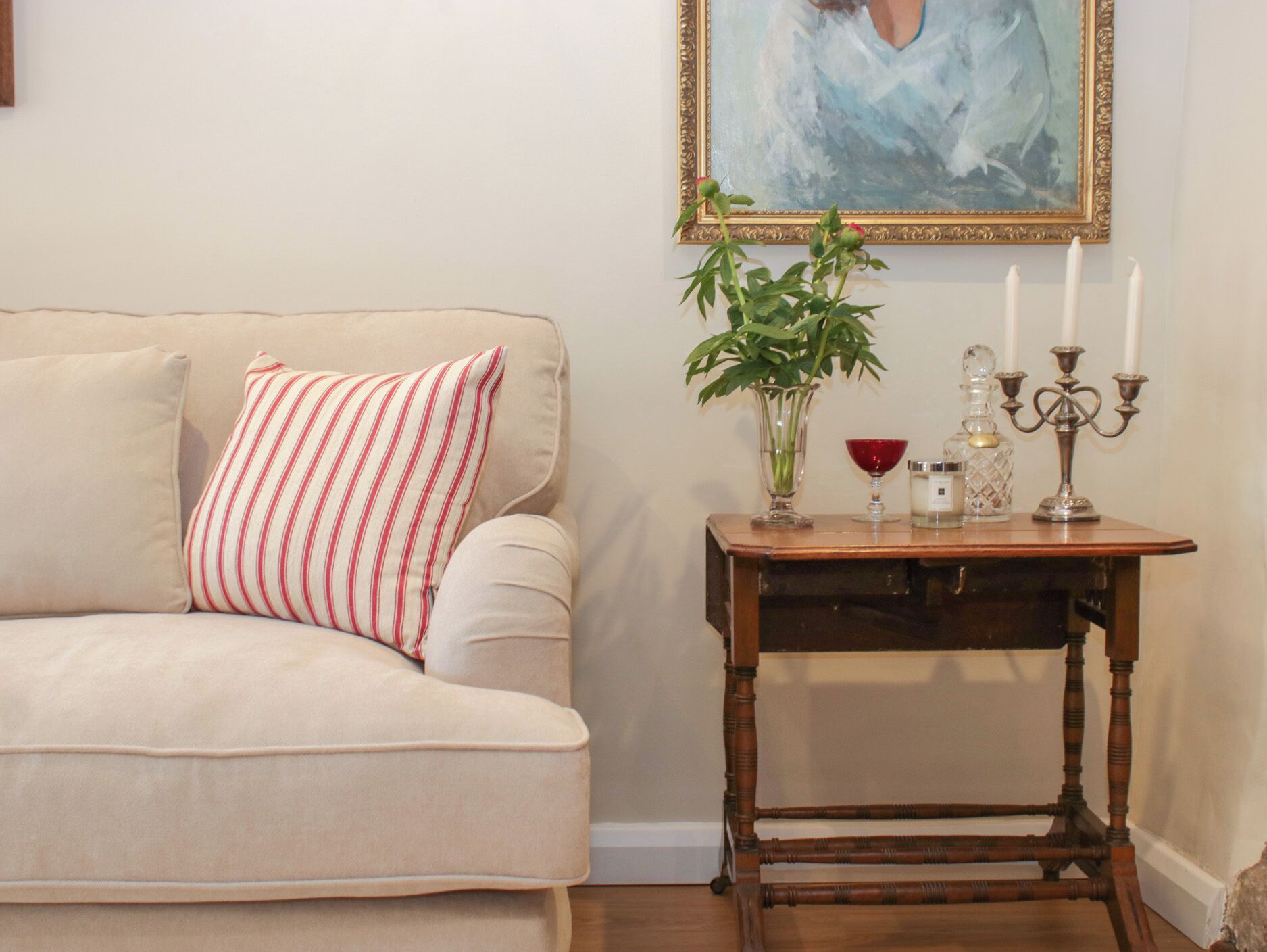 Un canapé beige agrémenté d’un coussin à rayures, une table d’appoint en bois sur laquelle sont posés un vase, des bougies et une carafe en verre.