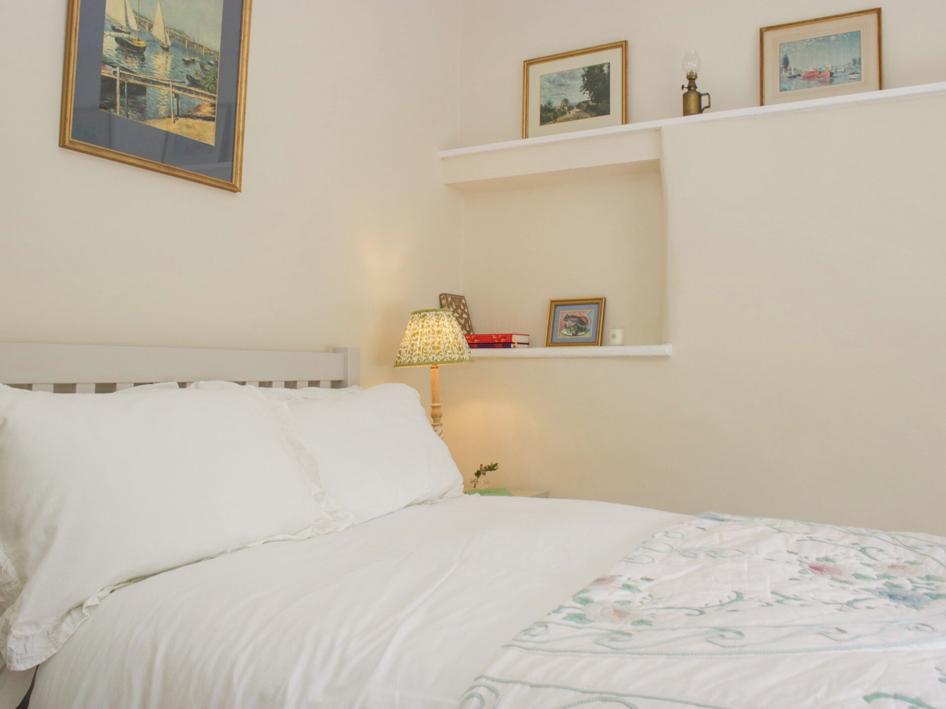 A neatly made bed with white linens, a bedside lamp, and a shelf with framed pictures.