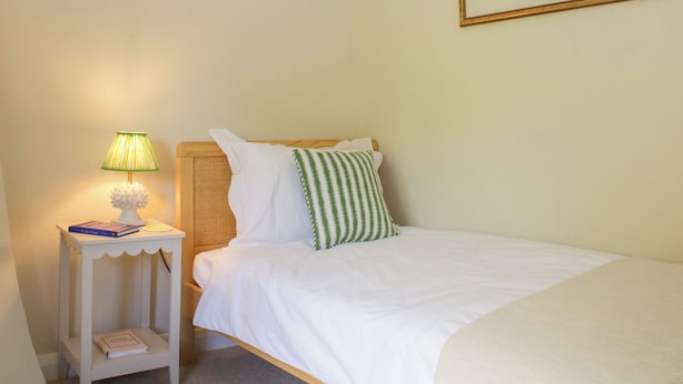 A bedroom with a bed, a bedside table with a lamp, a shelf with books and decorative items, and a framed painting on the wall. - Upper Frogpool Cottage (Crediton)