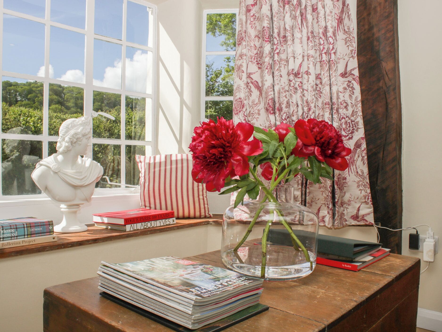 A room with a large window, a bust sculpture, a vase with flowers, and books on a wooden table.