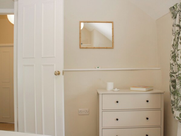 Ein Zimmer mit einer weißen Tür, einem Schminktisch mit Schubladen, einem Spiegel an der Wand und einem Regal über dem Schminktisch.