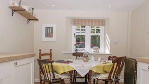 A dining area with a table set for four, wooden chairs, a radiator, a window with curtains, and a shelf above the table. - Upper Frogpool Cottage (Crediton)