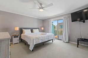 Ein Schlafzimmer mit Bett, Nachttischen, Deckenventilator, Fernseher und einem Balkon mit Vorhängen.