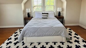 A bedroom with a large bed, two bedside tables with lamps, a window with blinds, and a patterned rug. - Hill Street Retreat (Oklahoma City)