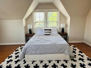 Ein Schlafzimmer mit einem großen Bett, zwei Nachttischen mit Lampen, einem Fenster mit Jalousien und einem gemusterten Teppich.