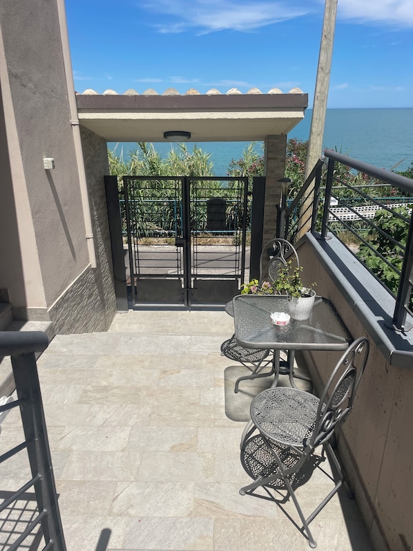 Un balcón con mesa y sillas, vista al mar y un portón que da a un jardín.