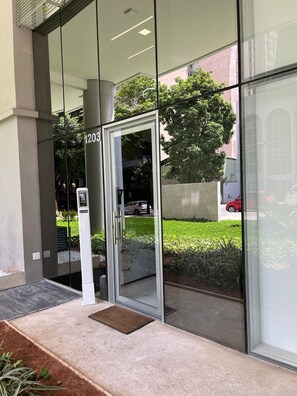 A modern building entrance with a glass door, a number '1203' sign, and a small outdoor area with plants. - LK Centro 10 (Belo Horizonte)