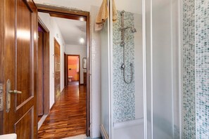 Un couloir avec des portes en bois, un espace douche orné de mosaïques et, au fond, une salle de bain visible.