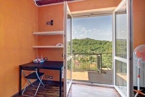 A room with a wooden floor, a black table with a chair, a window with a view of a green landscape, and a balcony with a railing.