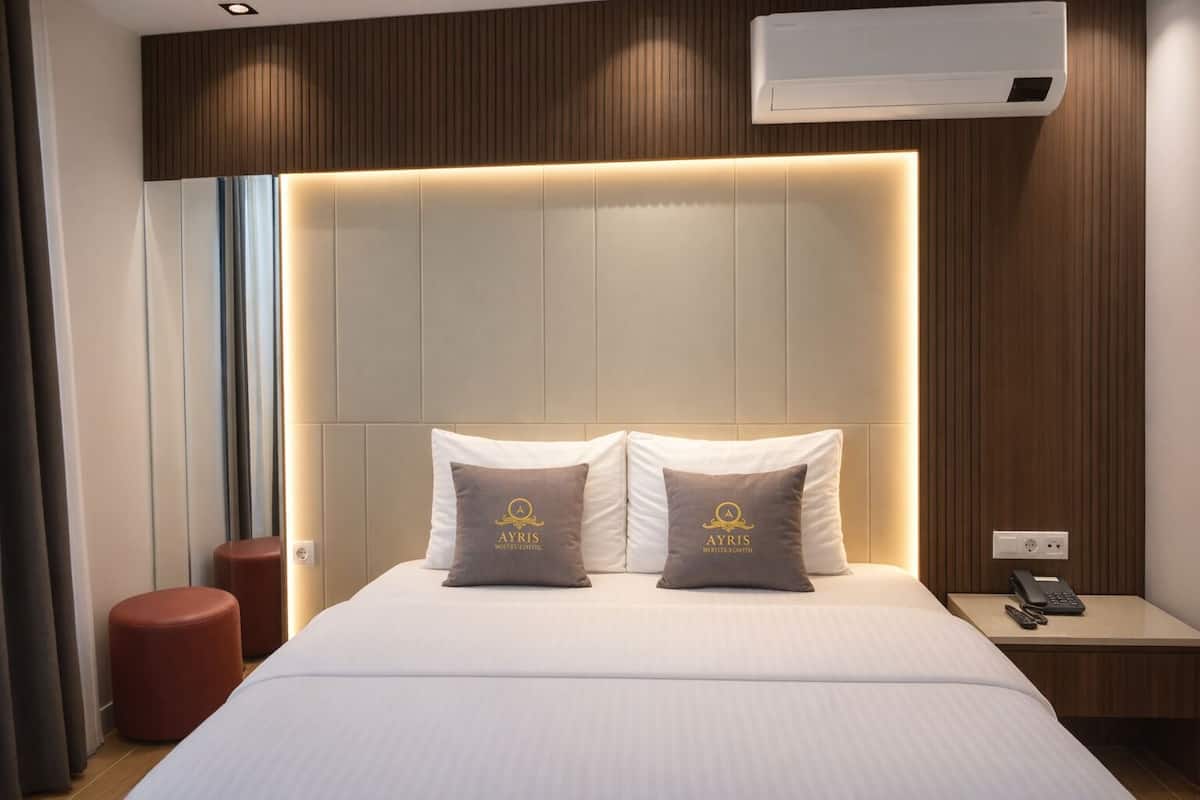 Una habitación de hotel con una cama, dos almohadas con un logotipo, una mesita de noche con un teléfono y un equipo de aire acondicionado.