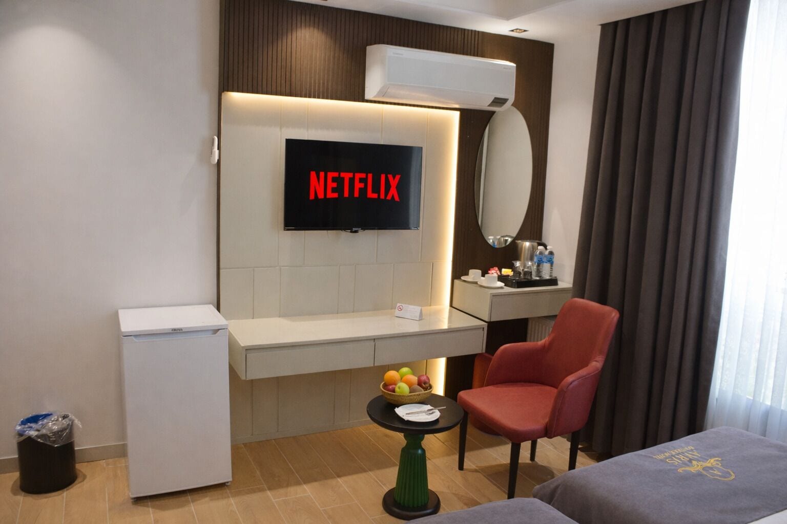 Et hotelværelse med et fjernsyn der viser 'NETFLIX', et lille bord med frugt, en rød stol, et minikøleskab og en seng.