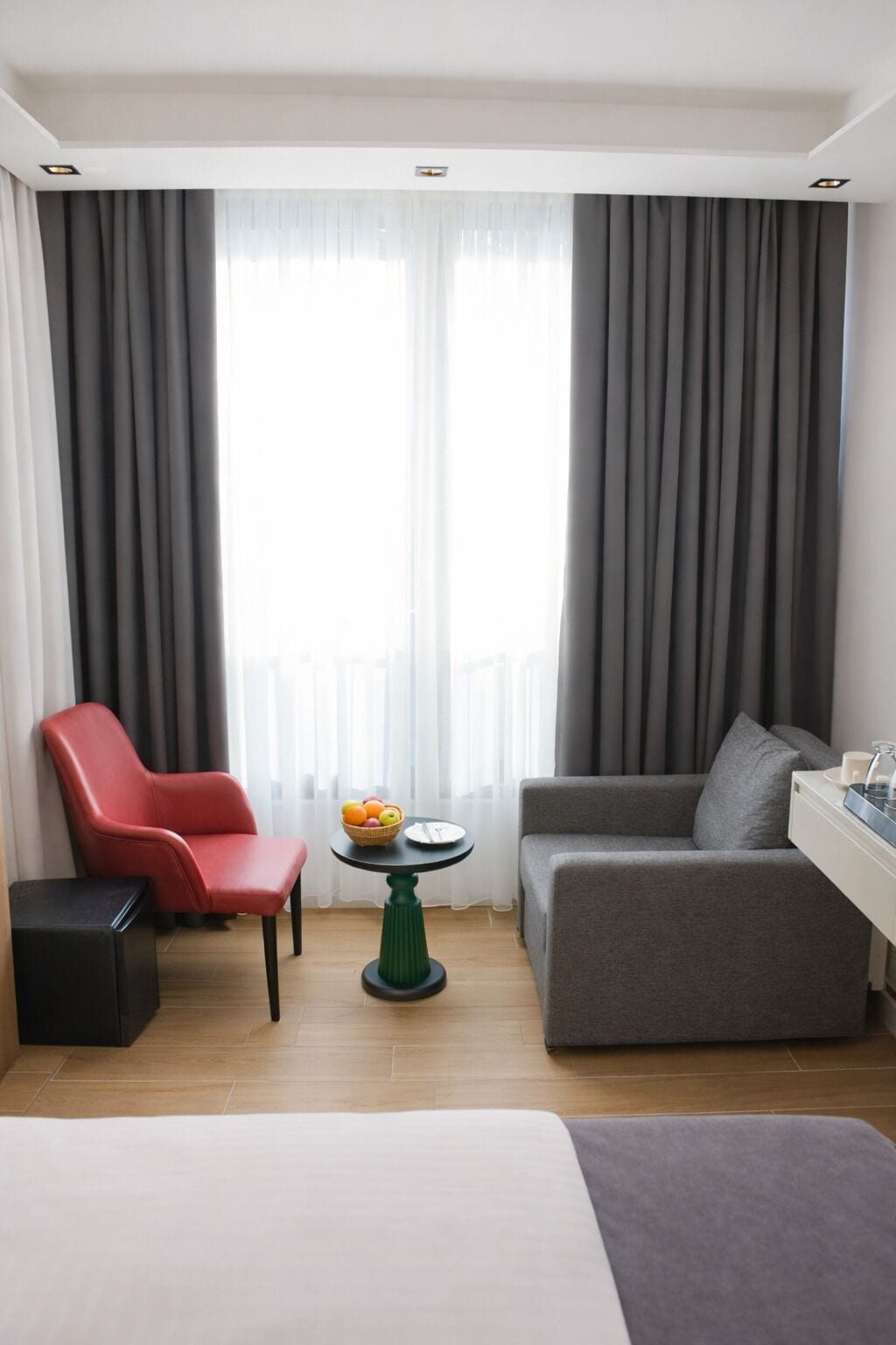 A hotel room with a bed, a red armchair, a grey sofa, a small round table with fruit, and a view through large windows with grey curtains.