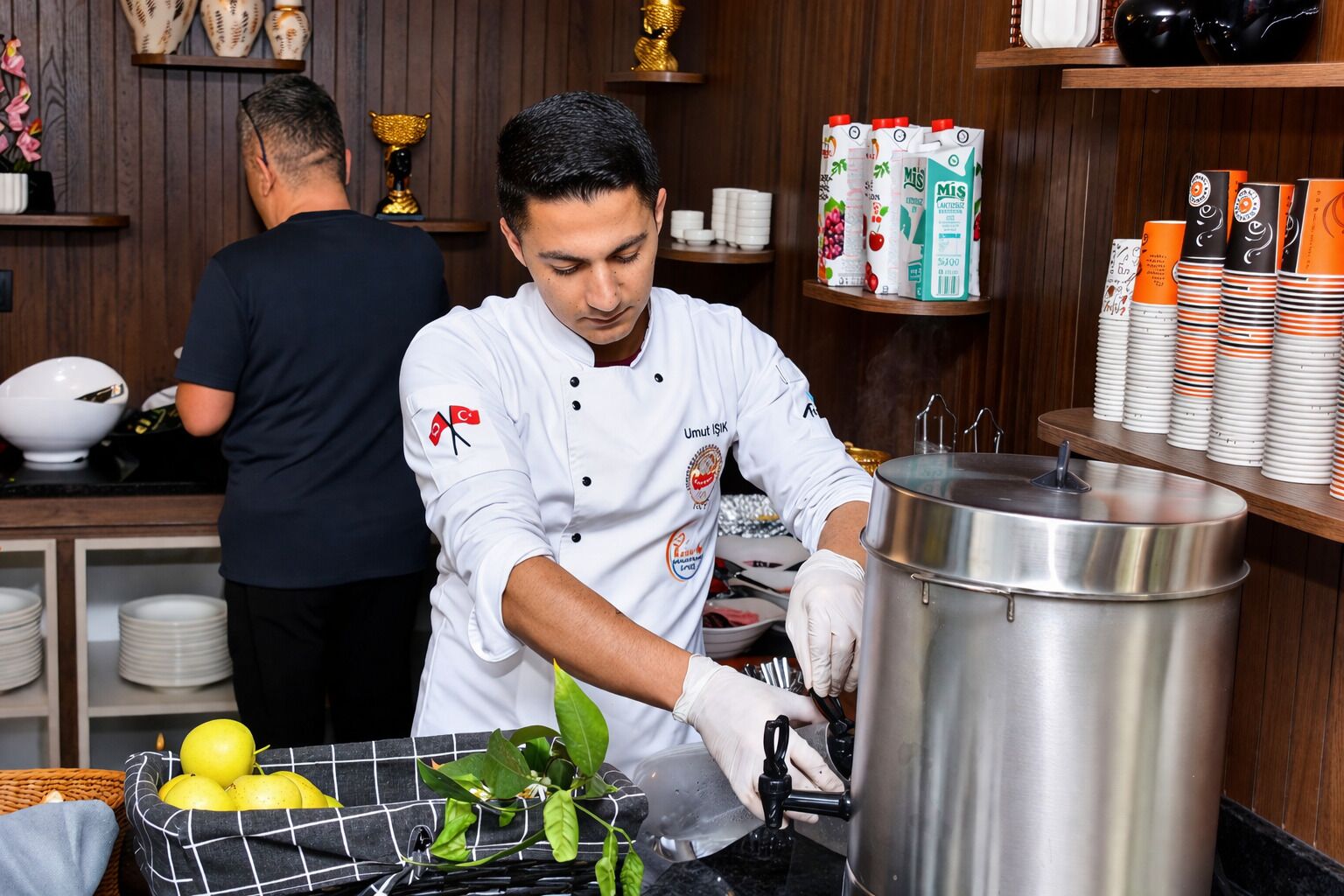 Un chef preparando comida en una cocina con una olla de acero inoxidable grande, un tazón de limones y una cesta de hojas verdes.