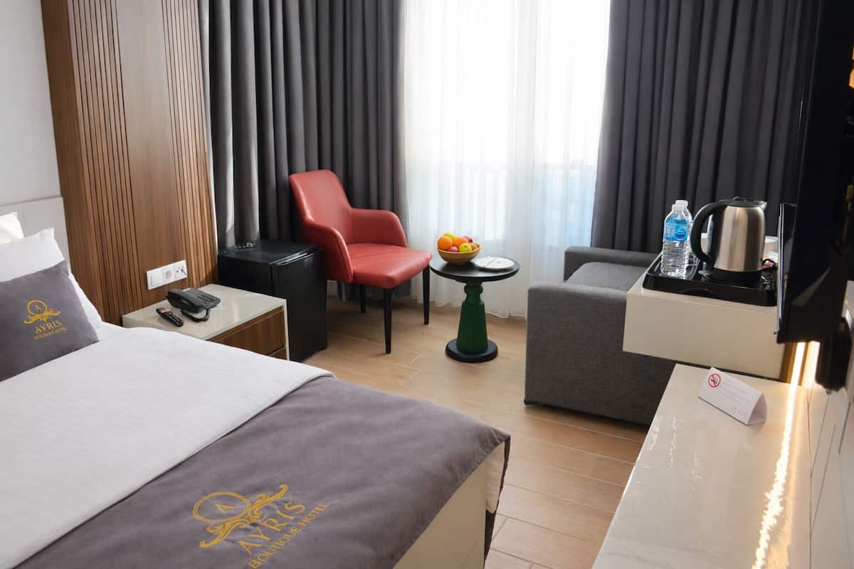 Habitación de hotel con cama, escritorio con teléfono, una silla roja, una mesita con fruta, un sillón gris y una cafetera.