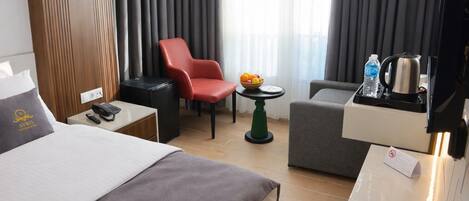 Une chambre d’hôtel avec un lit, un bureau avec un téléphone, une chaise rouge, une petite table avec des fruits, un fauteuil gris et une machine à café.