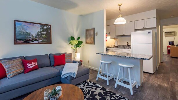 Una sala moderna con un sofá gris, una mesa de centro y una cocina con armarios blancos y encimera de mármol.