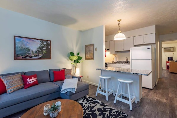 A modern living room with a gray sofa, a coffee table, and a kitchen area with white cabinets and a marble countertop. - Maple Row Apartment: Perfect for Game Day! (Bloomington)