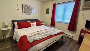Una cama bien hecha con ropa de cama roja y blanca, una mesita de noche con una lámpara, una ventana con cortinas rojas y un aire acondicionado instalado en la pared.
