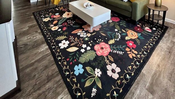 Un salon avec un tapis à motifs floraux, une table basse blanche, un canapé vert et deux lampes.