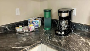 Une cuisine avec des meubles blancs, un plan de travail en marbre, une machine à café Keurig, une bouteille d’eau verte et une cafetière.