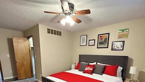 Une chambre à coucher avec un lit, un ventilateur de plafond, une table de chevet et des tableaux encadrés accrochés au mur.