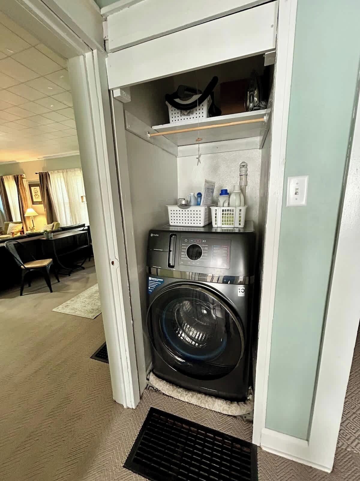 Une petite buanderie équipée d’un lave-linge et d’un sèche-linge à chargement frontal, d’étagères contenant des produits de lessive et d’un petit coin salon visible au fond.