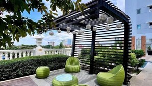 Een terras op het dak met groene zitplaatsen, een pergola met hangende lichtjes, en een uitzicht op gebouwen op de achtergrond.