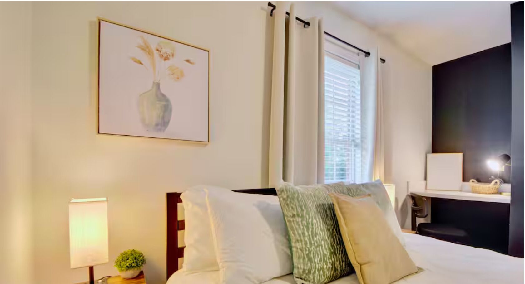Una habitación de hotel con cama, mesita de noche con lámpara, una planta pequeña, un cuadro en la pared y una ventana con cortinas.