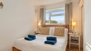 Uma cama bem arrumada com roupa de cama branca e toalhas azuis, cabeceira de madeira, duas luminárias de apoio e uma janela com cortinas.