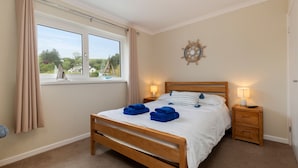 A bedroom with a large window, a wooden bed, two bedside tables with lamps, and a wall decoration. - Smugglers Cove - Short Walk to Beach (Amroth)