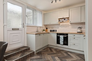A kitchen with white cabinets, a herringbone wood floor, a built-in oven, and a window with blinds. - Smugglers Cove - Short Walk to Beach (Amroth)