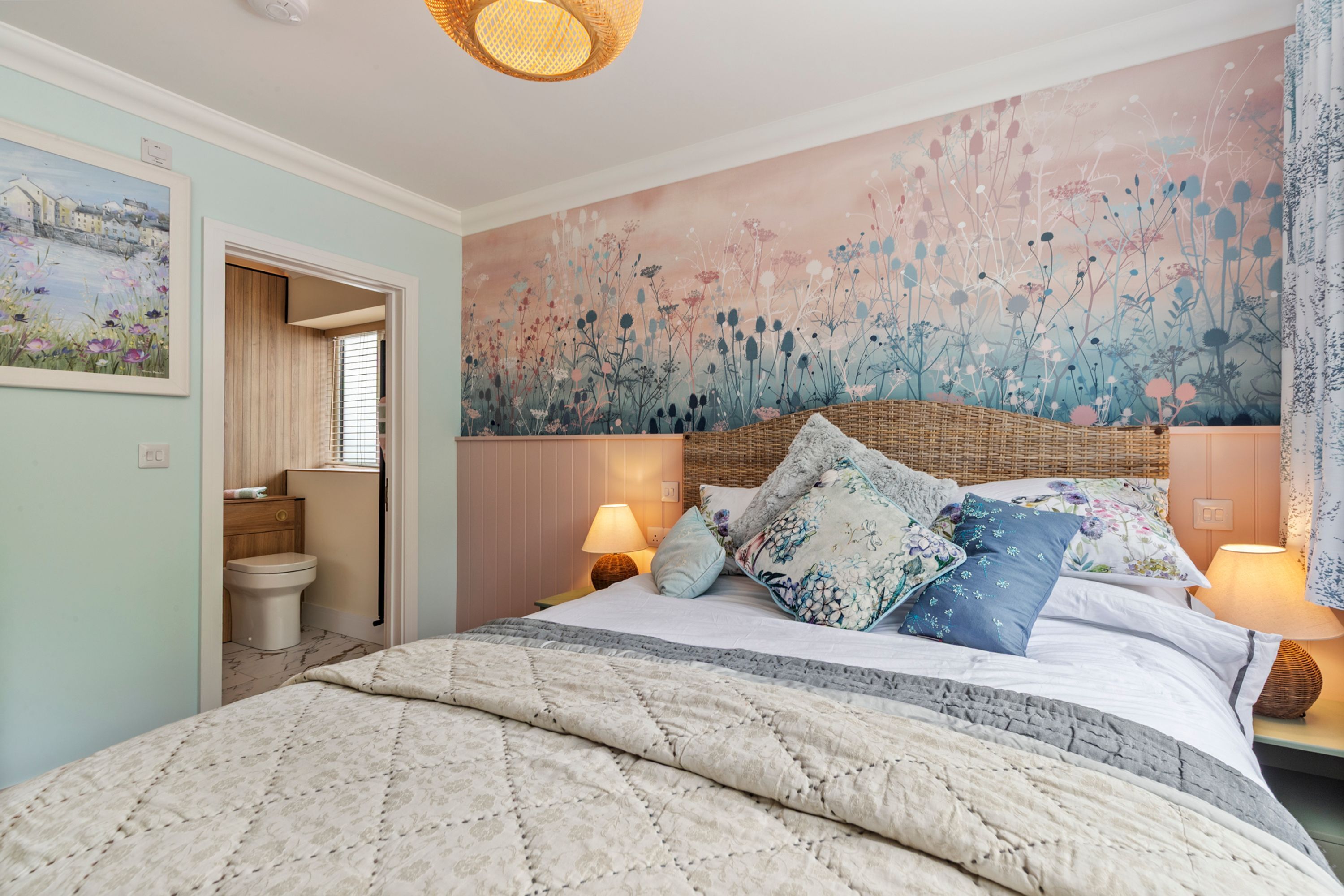 A bedroom with a bed, two bedside lamps, a wall mural, and a door leading to a bathroom.