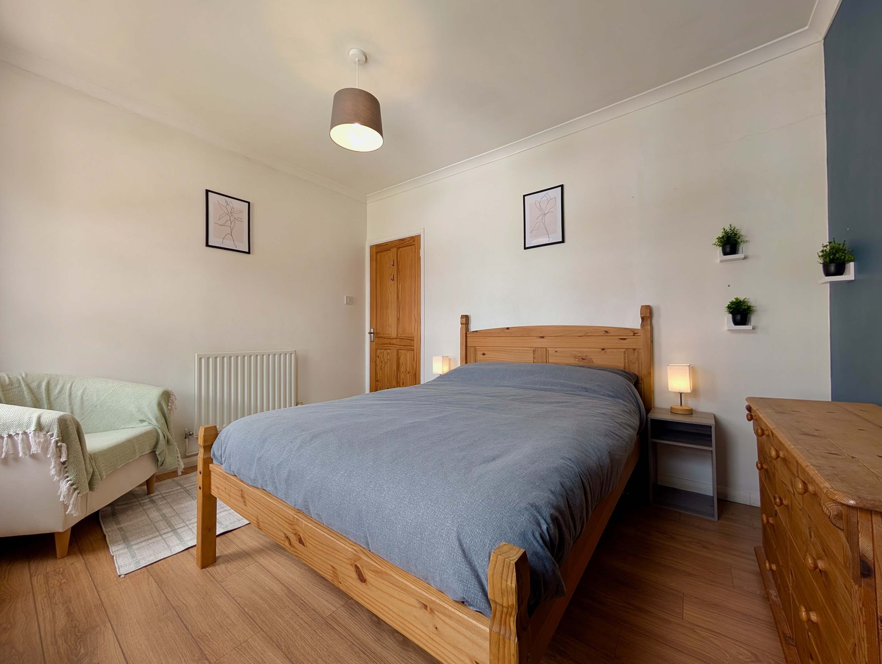 A bedroom with a wooden bed, a bedside table with a lamp, a radiator, a door, and a small shelf with plants.